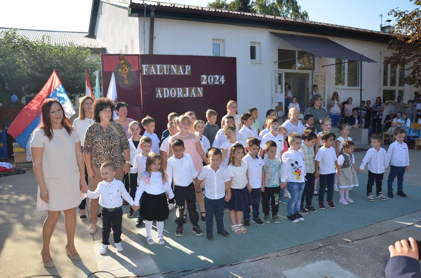 Gyerekek és pedagógusok fellépése az adorjáni falunapon, a háttérben Falunap 2024 Adorján felirat