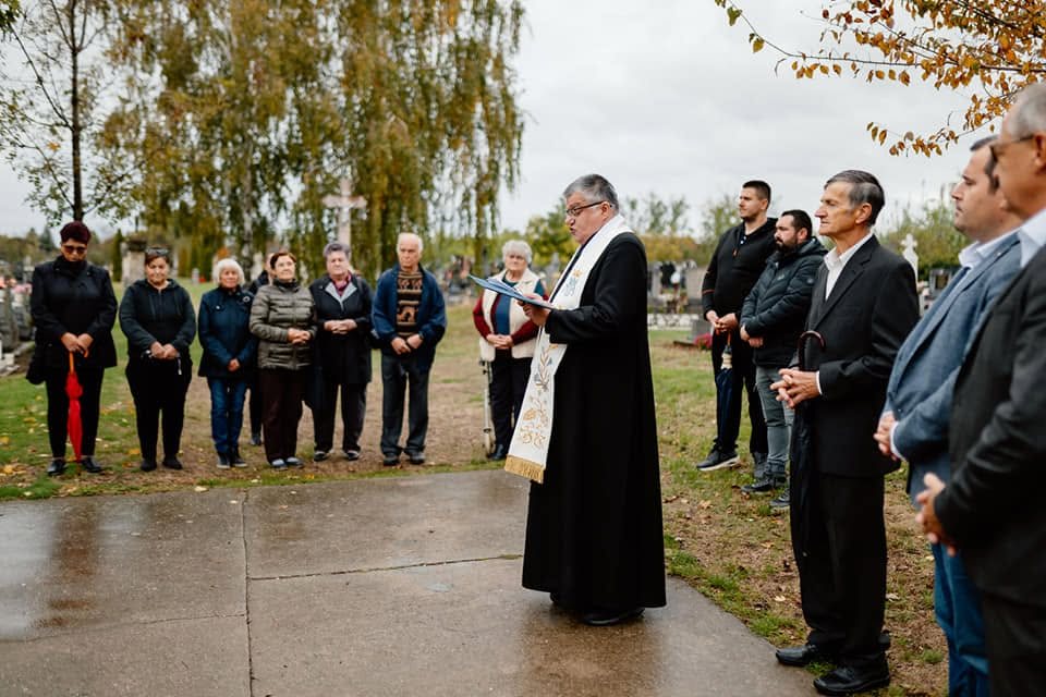 Szabadtéri megemlékezés egy temetőben, pap mond imát, körülötte helyiek és a község képviselői